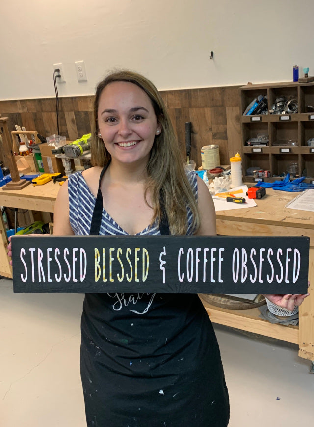 A person holding a wooden sign with the text 'STRESSED BLESSED & COFFEE OBSESSED' on it.