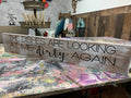 A wooden single plank sign with the humorous text 'THE DISHES ARE LOOKING AT ME DIRTY AGAIN' in capital letters.