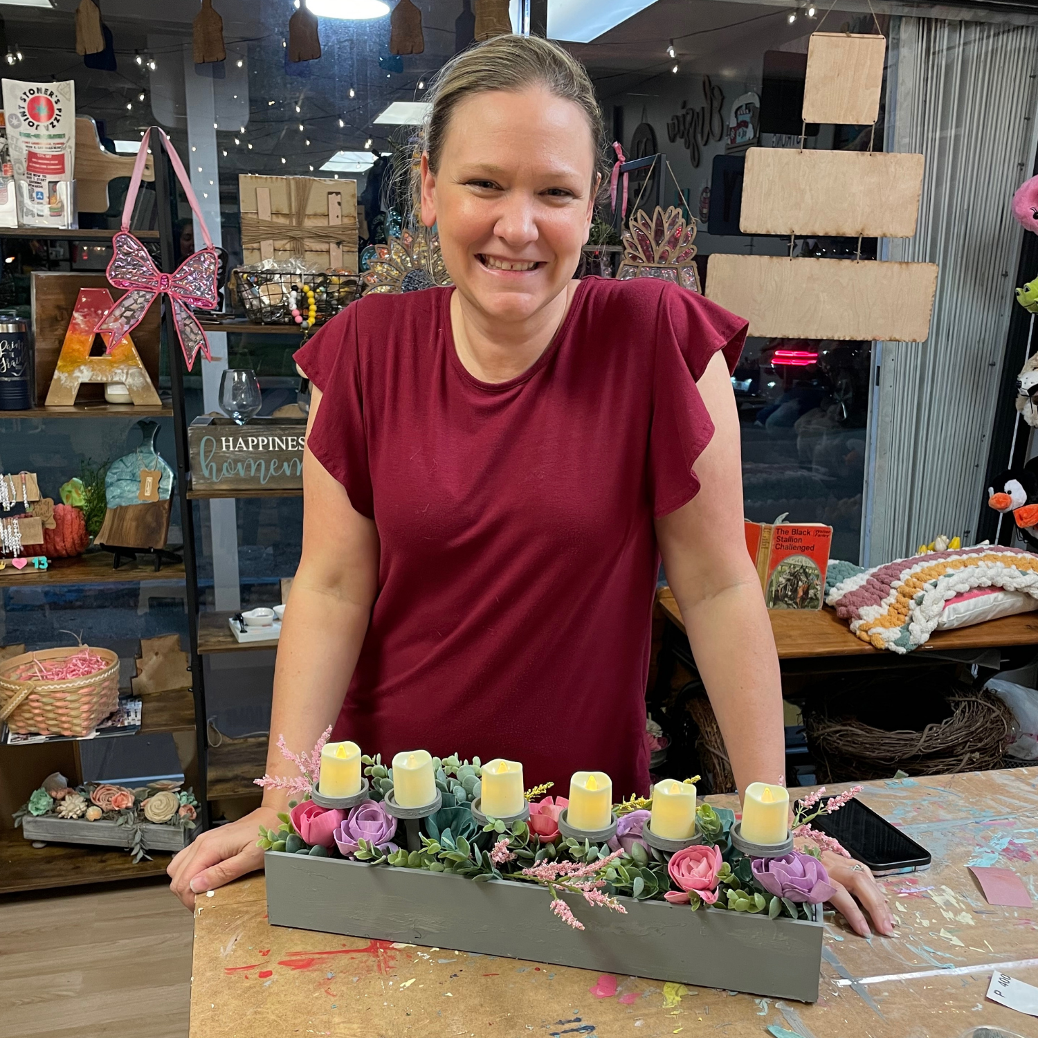 Woman in a red shirt standing behind a table with a floral and candle arrangement in a store setting.