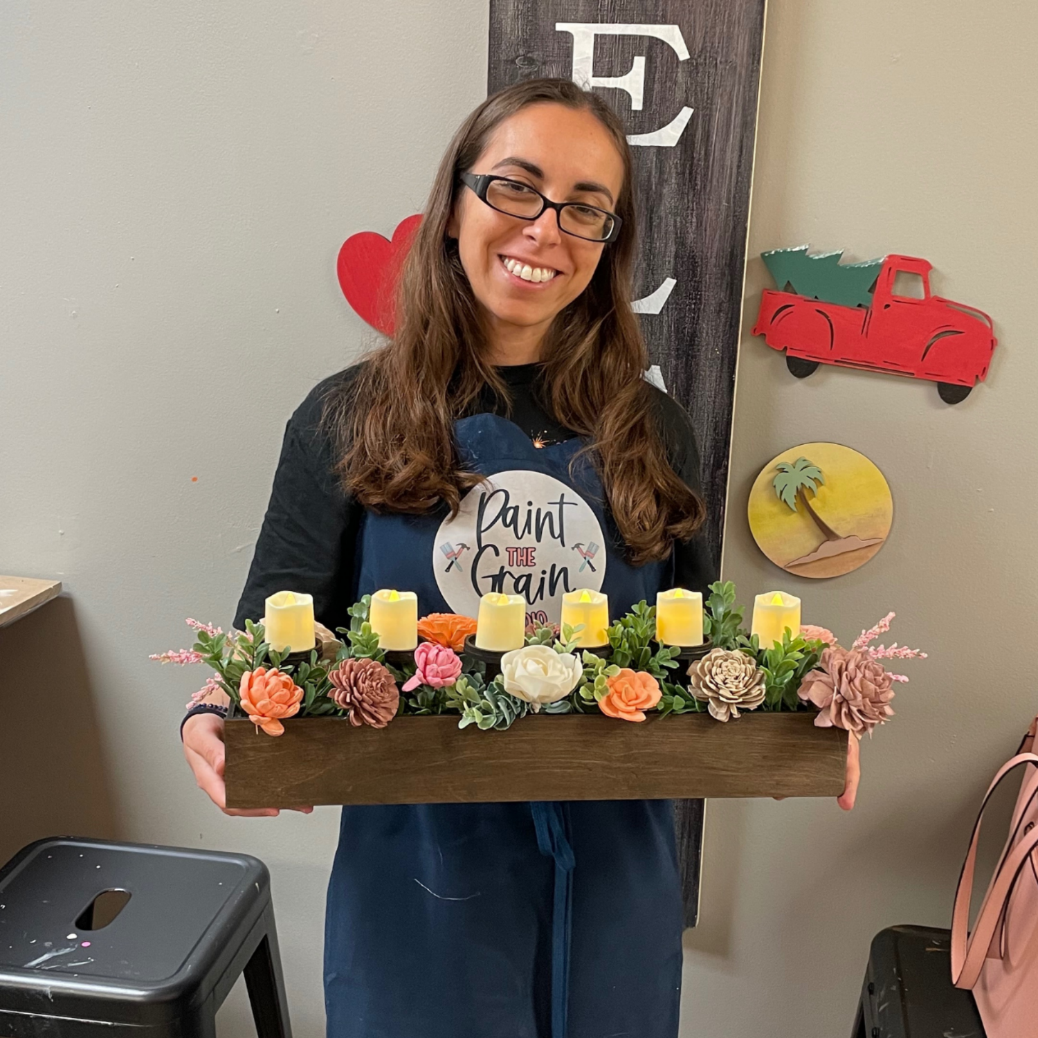 Person holding a wooden tray with candles and flowers against a wall with decorative items.