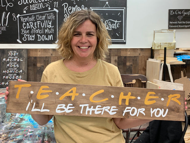 A person holding a wooden sign with the word 'TEACHER' followed by the phrase 'I'll be there for you' in stylized lettering.