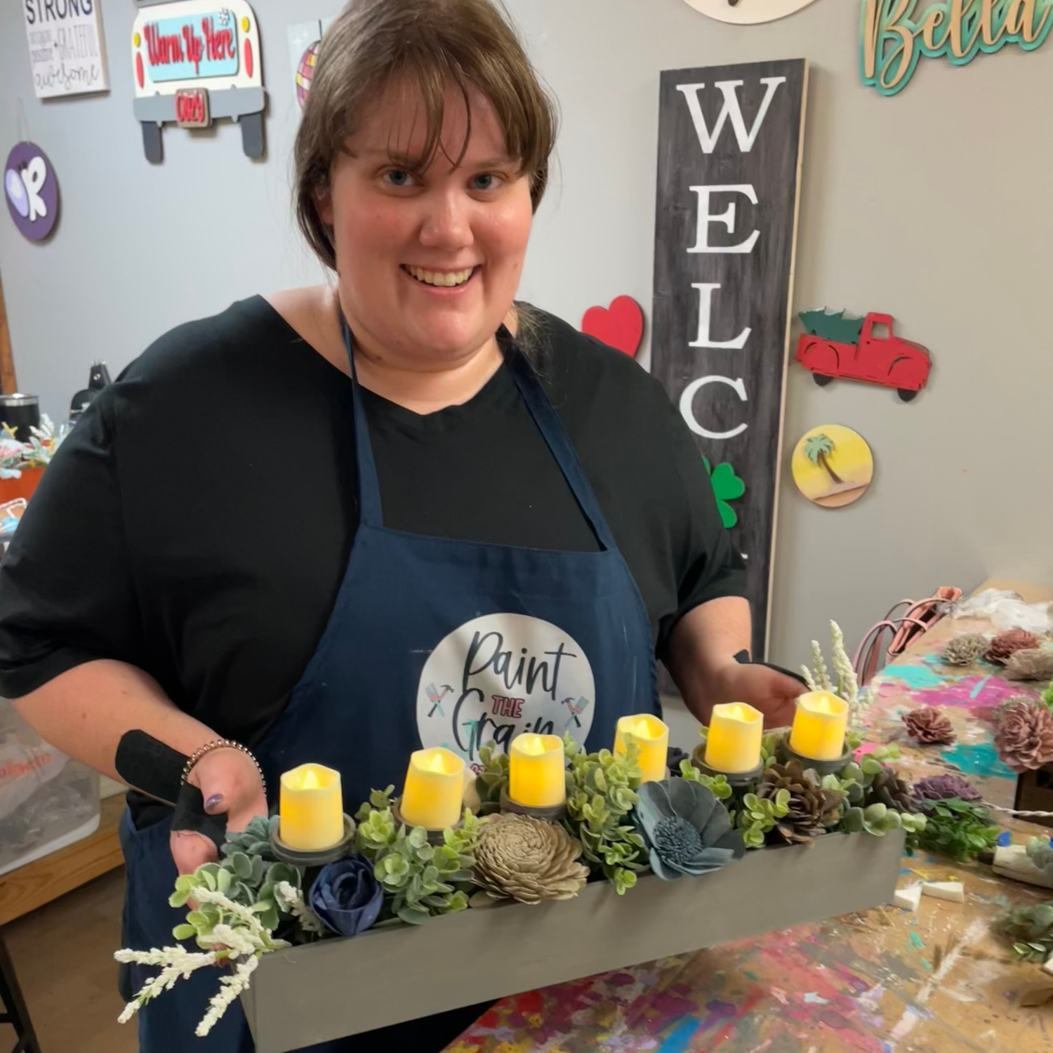 Person wearing a blue apron holding a decorative tray with candles and flowers in a craft or painting studio.